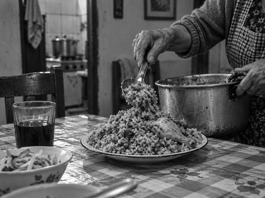 Montaña de arroz con pollo en un plato, manos de una señora mayor sirviendo más, vaso de chicha y sarsa sobre mantel de hule.