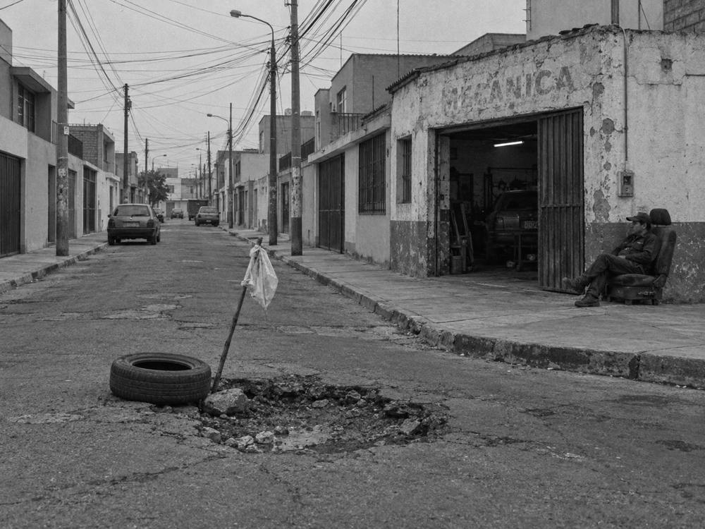 Bache señalizado con una llanta vieja y un palo con una bolsa amarrada; al fondo, el mecánico sentado frente a su taller.