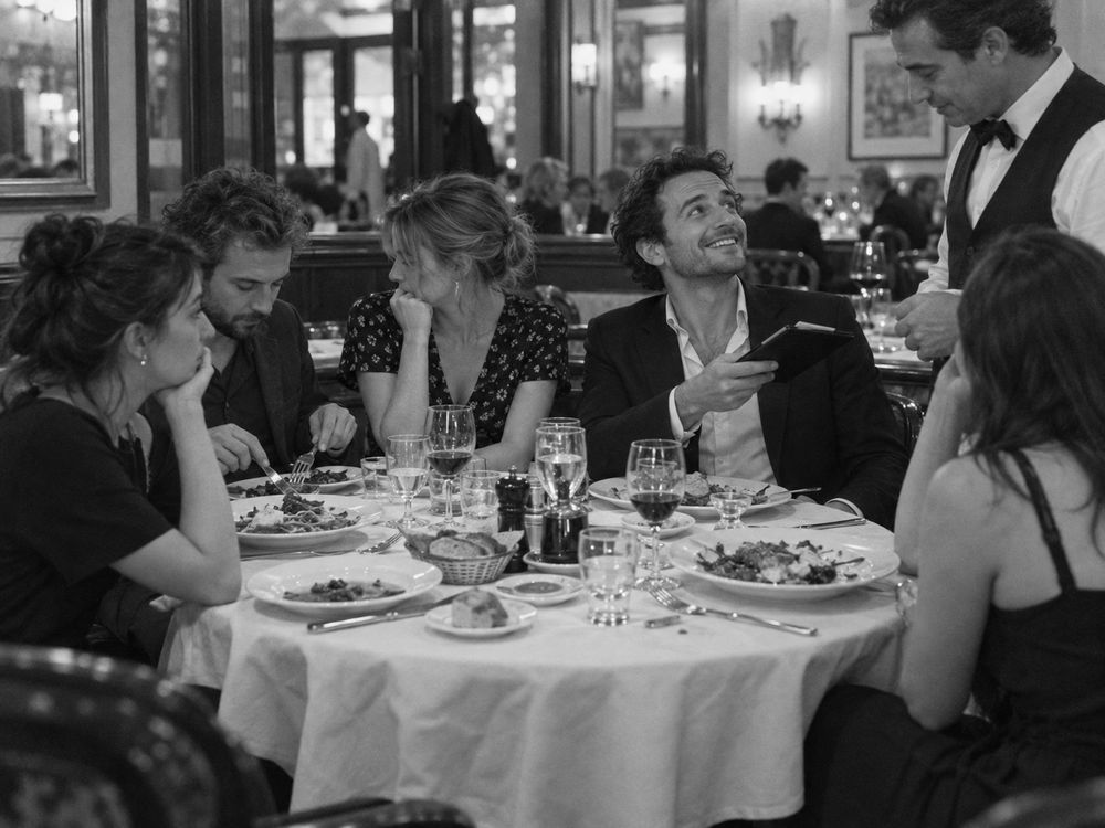 Mesa de restaurante con platos variados; un comensal sonriente sostiene la cuenta mientras otro mira su ensalada.