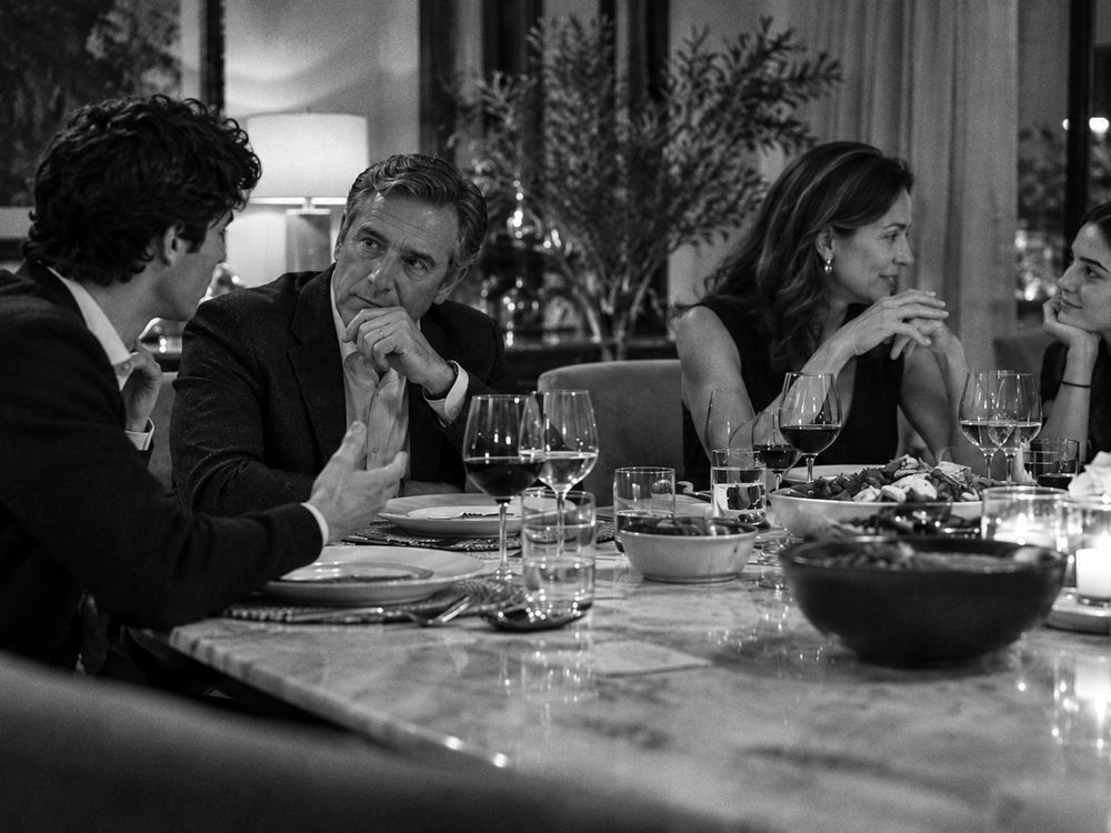 Cena familiar en un comedor elegante; un hombre joven pregunta algo y el padre lo mira por encima de los lentes.