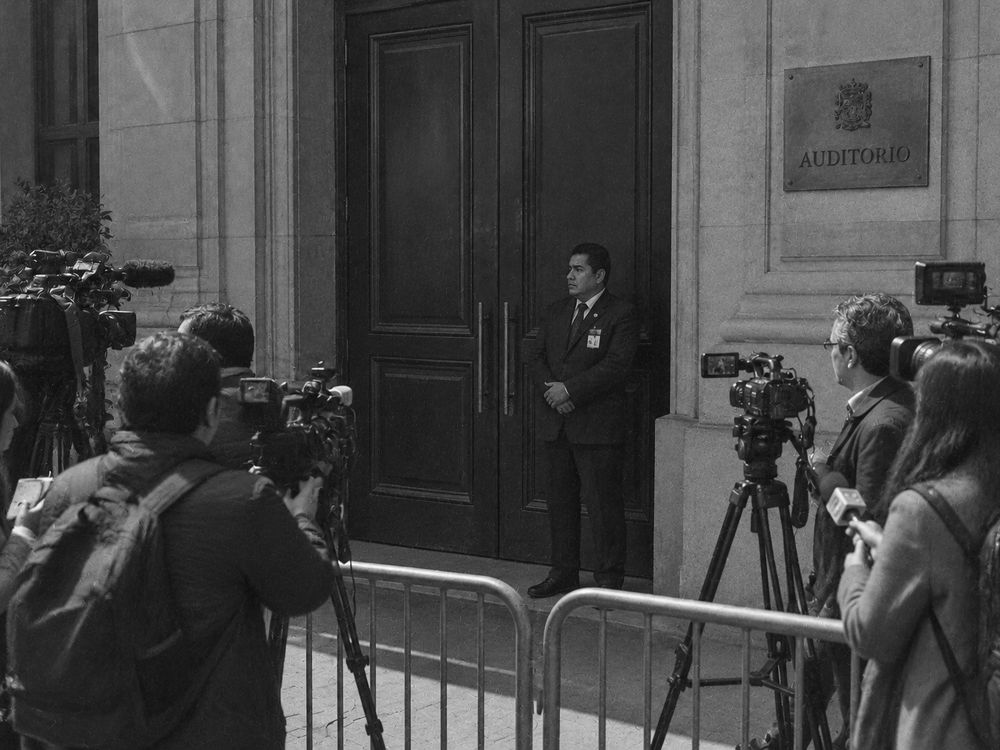 Puerta cerrada de un auditorio institucional, con un vigilante delante y periodistas esperando afuera.