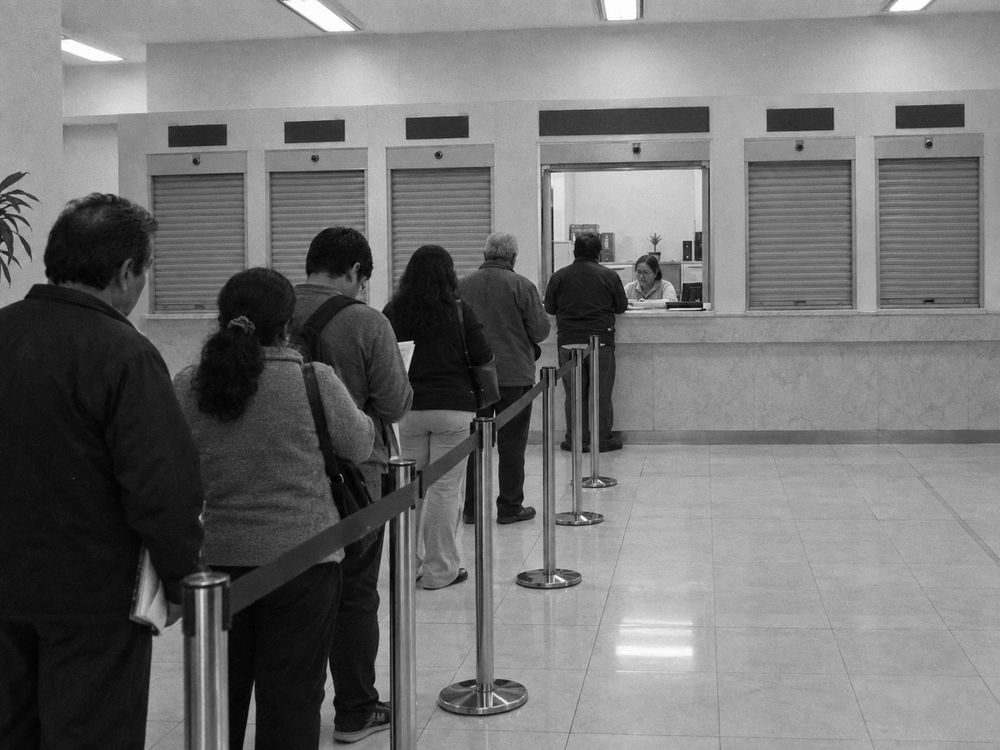 Módulo de atención con siete ventanillas, cuatro con la cortina bajada, y una fila de personas frente a una sola ventanilla abierta.