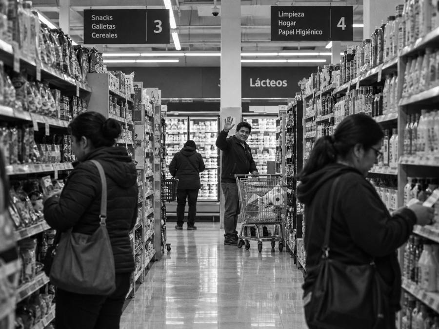 Un hombre con su carrito saluda a cámara desde el fondo del pasillo de un supermercado, sin acercarse.